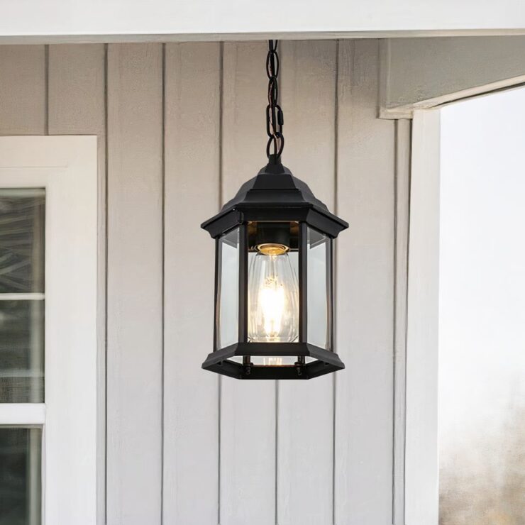 Un lustre extérieur pour terrasse de style contemporain en métal noir, de forme hexagonale, avec des panneaux de verre transparents entourant une ampoule centrale. Il est fixé au plafond par une chaîne en métal. Le lustre est installé sur un porche en bois, ajoutant une touche élégante à l'extérieur de la maison.