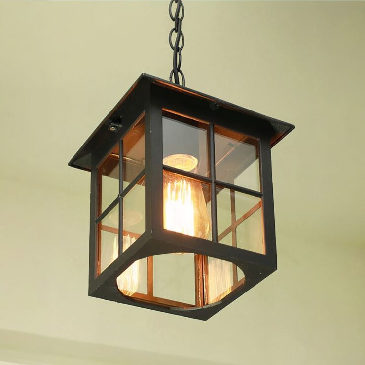Un lustre extérieur pour terrasse de style vintage est suspendu avec une structure carrée en métal noir. Les côtés sont composés de vitres transparentes encadrées, permettant de voir une ampoule à filament à l'intérieur. Le luminaire est suspendu par une chaîne noire.