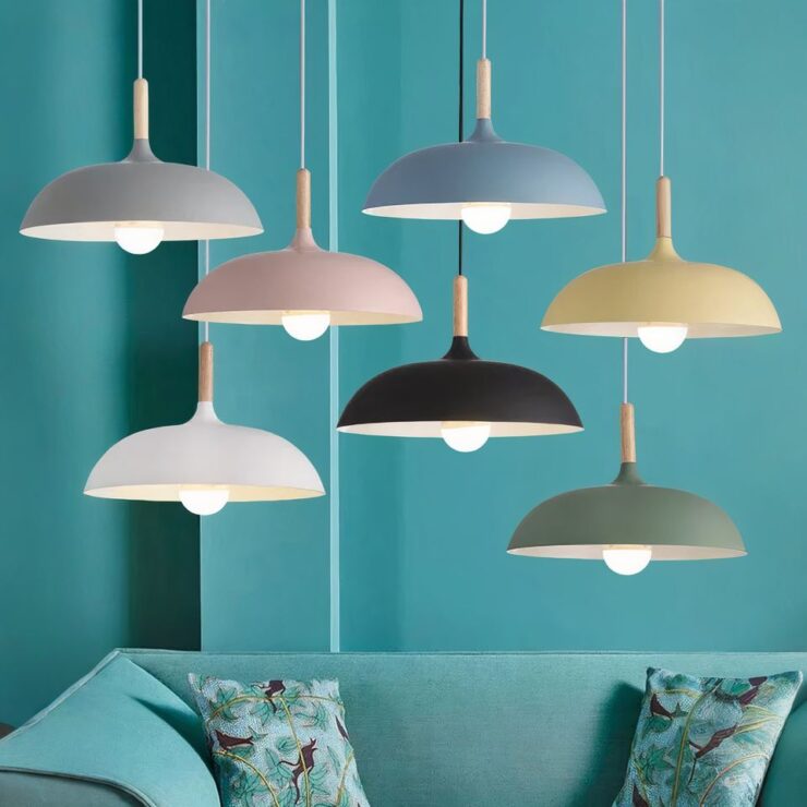 Sept lustres scandinave ronds en métal et bois, dans des couleurs variées telles que le gris, le rose, le bleu, le noir, le blanc, vert et jaune. Chaque suspension a une tige en bois et est suspendue dans un intérieur avec un mur bleu et un canapé assorti.