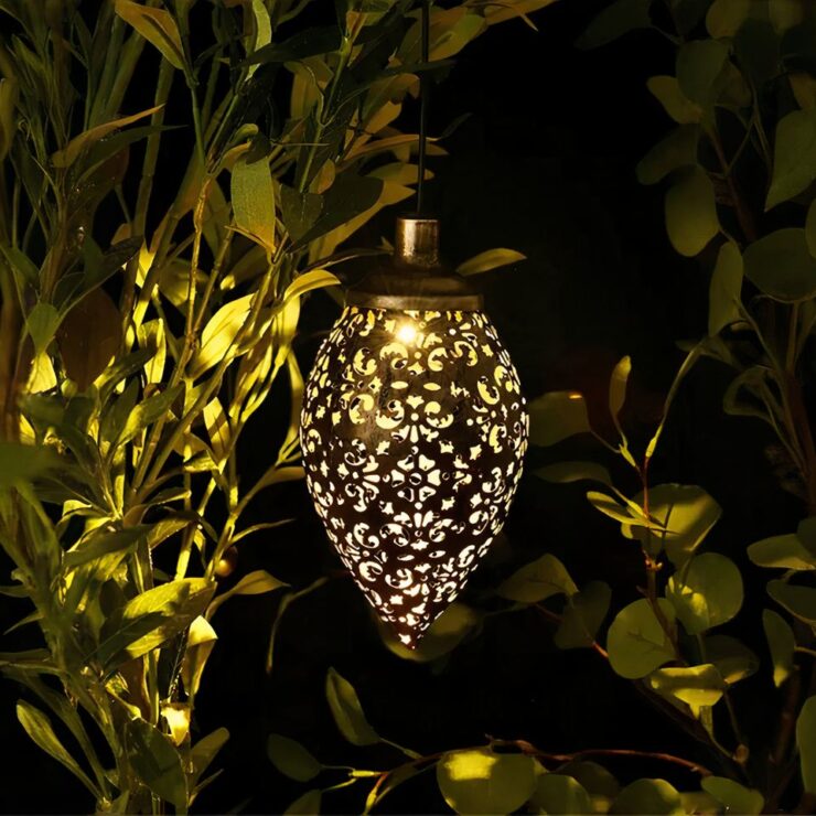 Lustre extérieur solaire en forme de goutte d'eau avec un design ajouré, suspendu dans un jardin, projetant des motifs lumineux complexes sur les feuilles et les environs, créant une ambiance chaleureuse et apaisante.
