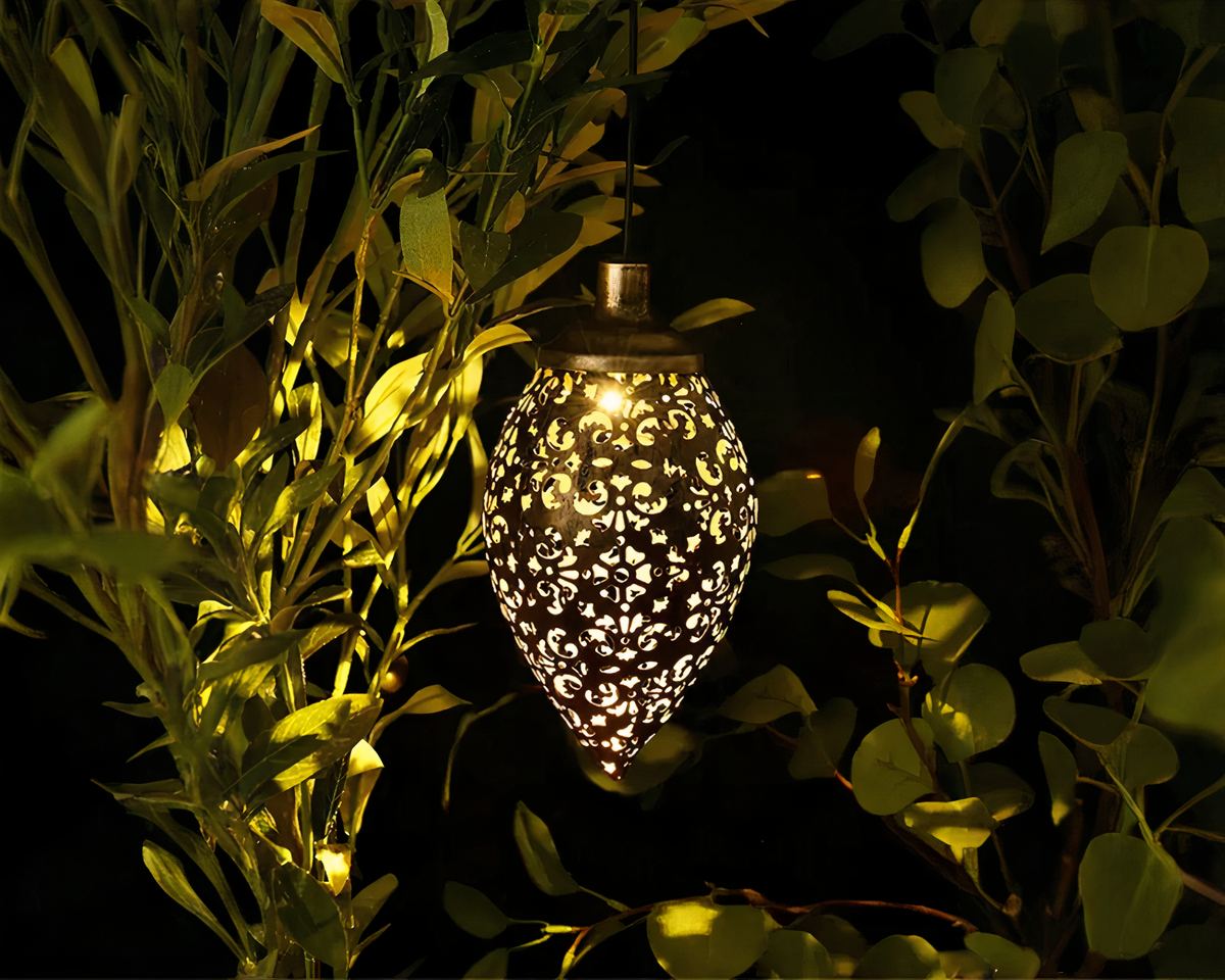 Lustre extérieur solaire en forme de goutte d'eau avec un design ajouré, suspendu dans un jardin, projetant des motifs lumineux complexes sur les feuilles et les environs, créant une ambiance chaleureuse et apaisante.