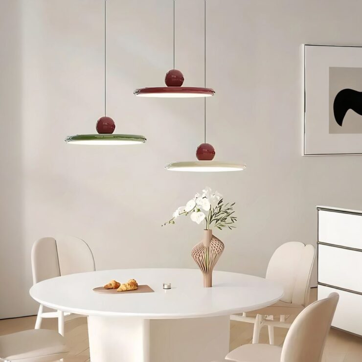 Trois lustres vintage en forme de disque sont suspendus au-dessus d'une table ronde blanche. Chaque lustre a une boule rouge au sommet, ils sont de différentes couleurs : l'un est rouge, l'autre vert et le troisième blanc. La lumière est douce et crée une ambiance chaleureuse dans une salle à manger moderne.