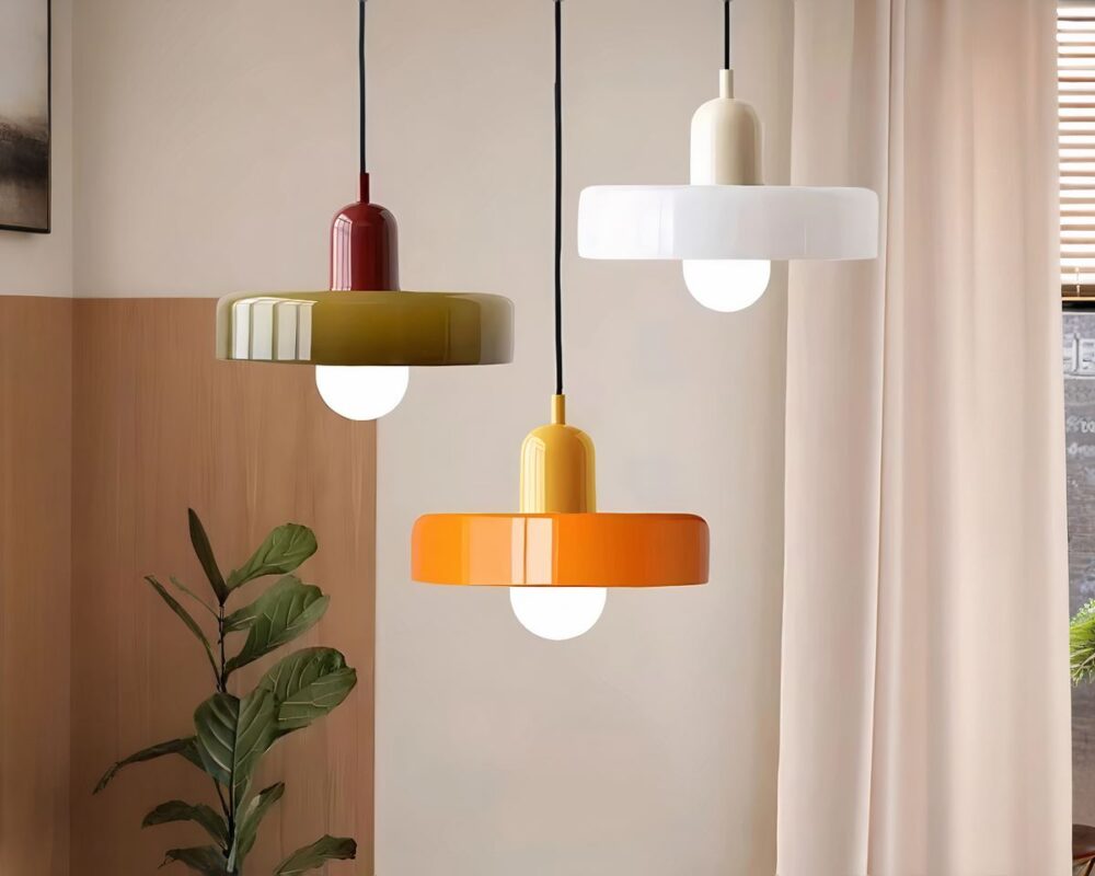 Trois lustres vintage ronds en verre coloré, aux teintes blanche, orange et blanc, suspendus au plafond dans une pièce minimaliste avec une plante verte en arrière-plan.