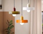 Trois lustres vintage ronds en verre coloré, aux teintes blanche, orange et blanc, suspendus au plafond dans une pièce minimaliste avec une plante verte en arrière-plan.