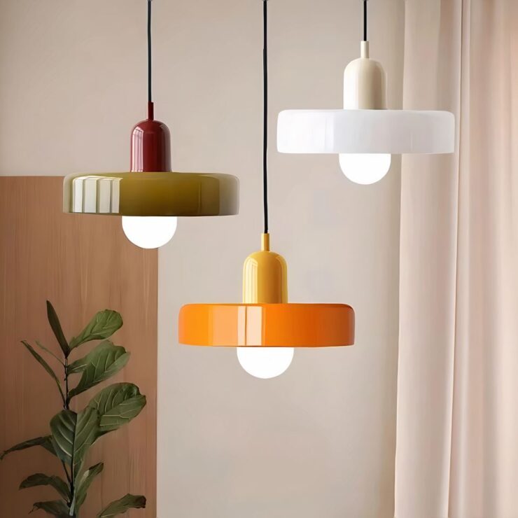 Trois lustres vintage ronds en verre coloré, aux teintes blanche, orange et blanc, suspendus au plafond dans une pièce minimaliste avec une plante verte en arrière-plan.
