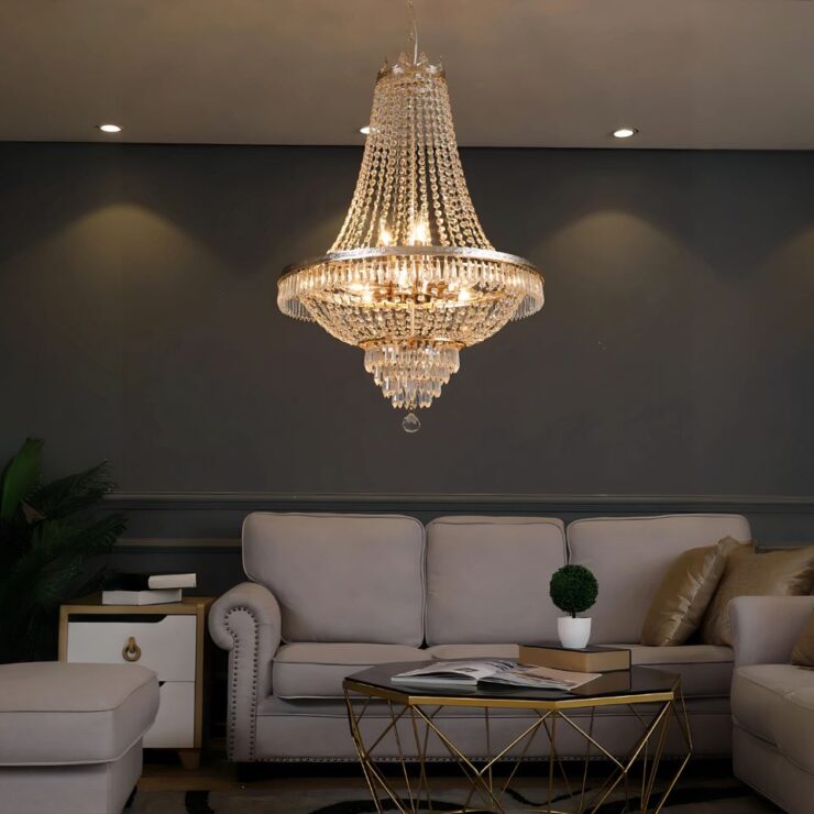 On voit une pièce aux murs gris foncés et un plafond blanc avec des LED encastrées le long du mur. Contre le mur, un canapé 3 places de couleur grise, un fauteuil assorti à gauche et à droite. Au centre une table basse, avec les pieds en fer et une petite plante verte dessus. Au plafond, un lustre chic et luxueux effet cristal dont la lumière est blanc chaud. La structure du lustre est argentée.
