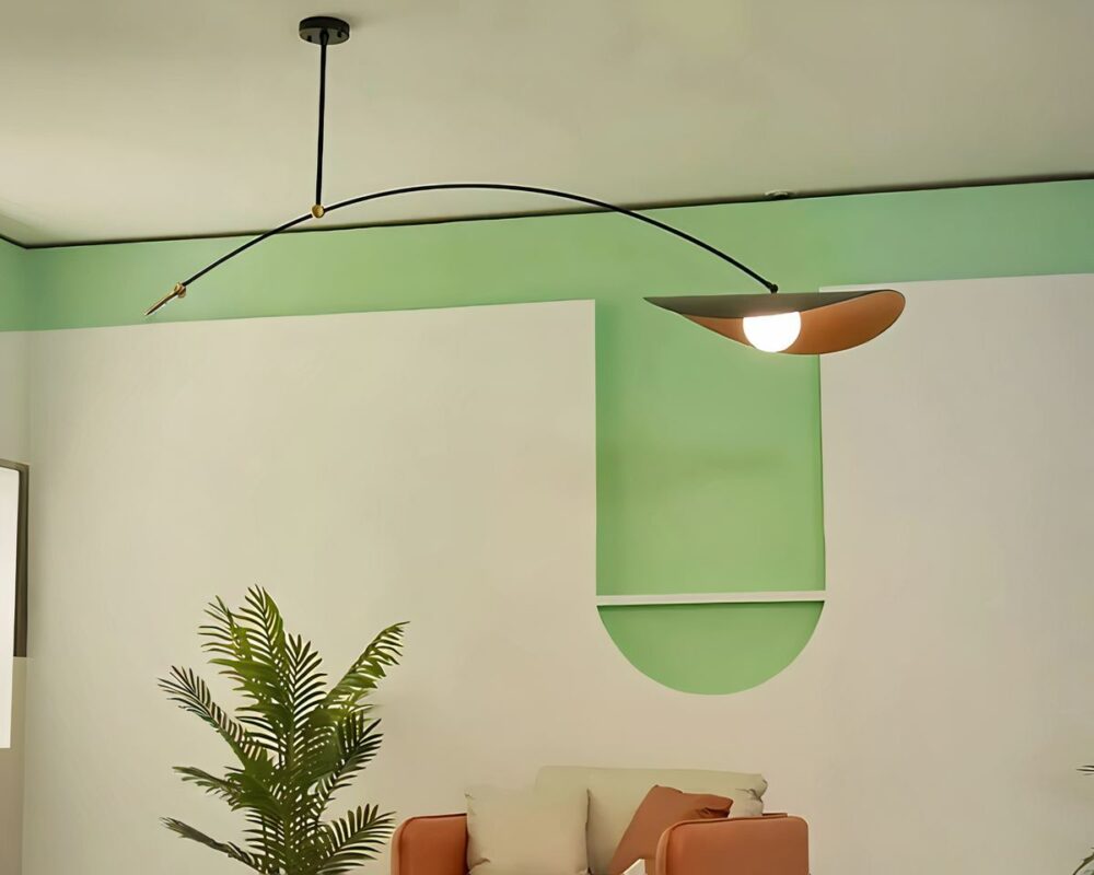 Lustre long avec bras suspendu fixé sur un plafond beige. Il est composé d'un socle rond noir au plafond, d'où descend une tige fine noire en métal. A l'extrémité de cette tige, un bras fin noir courbé vers le bras avec à droite un abat-jour rond avec les bords courbés vers le bas. L'extérieur est noir et l'intérieur est doré. L'ampoule est allumée. Le mur derrière est beige et vert clair. On voit contre le mur, une plante verte et un canapé beige et marron.