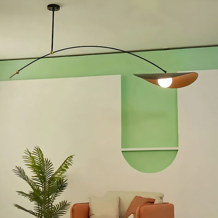 Lustre long avec bras suspendu fixé sur un plafond beige. Il est composé d'un socle rond noir au plafond, d'où descend une tige fine noire en métal. A l'extrémité de cette tige, un bras fin noir courbé vers le bras avec à droite un abat-jour rond avec les bords courbés vers le bas. L'extérieur est noir et l'intérieur est doré. L'ampoule est allumée. Le mur derrière est beige et vert clair. On voit contre le mur, une plante verte et un canapé beige et marron.