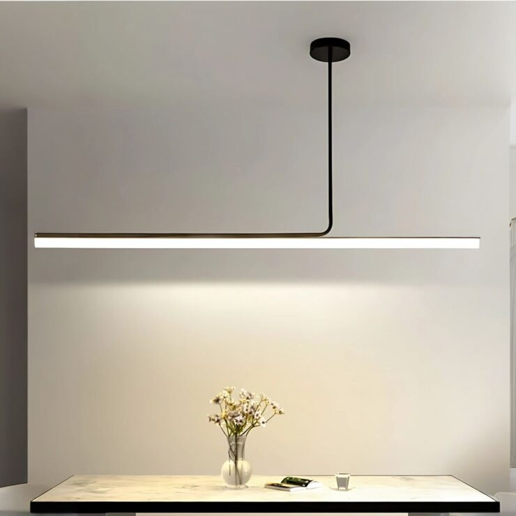 Lustre long fin et minimaliste fixé sur un plafond blanc. Un mur blanc à l'arrière. Le lustre est composé d'une tige noir verticale et d'une partie longue horizontale lumineuse. Sous le lustre une table avec un petit vase avec des fleurs.