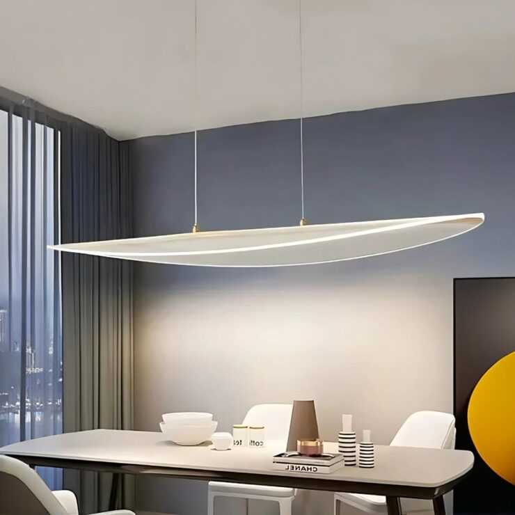 Lustre long suspendu en acrylique de forme ovale au-dessus d'une table blanche avec des chaises assorties. A gauche, on voit une grande fenêtre avec un rideau gris. Le mur derrière la table est gris. Un tableau noir et orange sur la droite du mur. Sur la table, de la vaisselle blanche et des livres.
