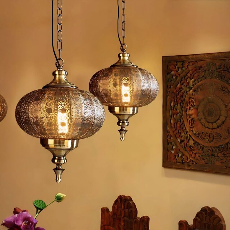 On voit deux lustres marocains en métal doré ajouré laissant passer la lumière blanc chaud donnant une ambiance lumineuse orangée. Ils sont suspendus par une chaine assortie et ont une forme ronde avec un style goutte sur le bas. Sur le mur de droite, un tableau style carré en bois sculpté. On perçoit deux chaises en bois sur le bas de la photo et une plante verte.
