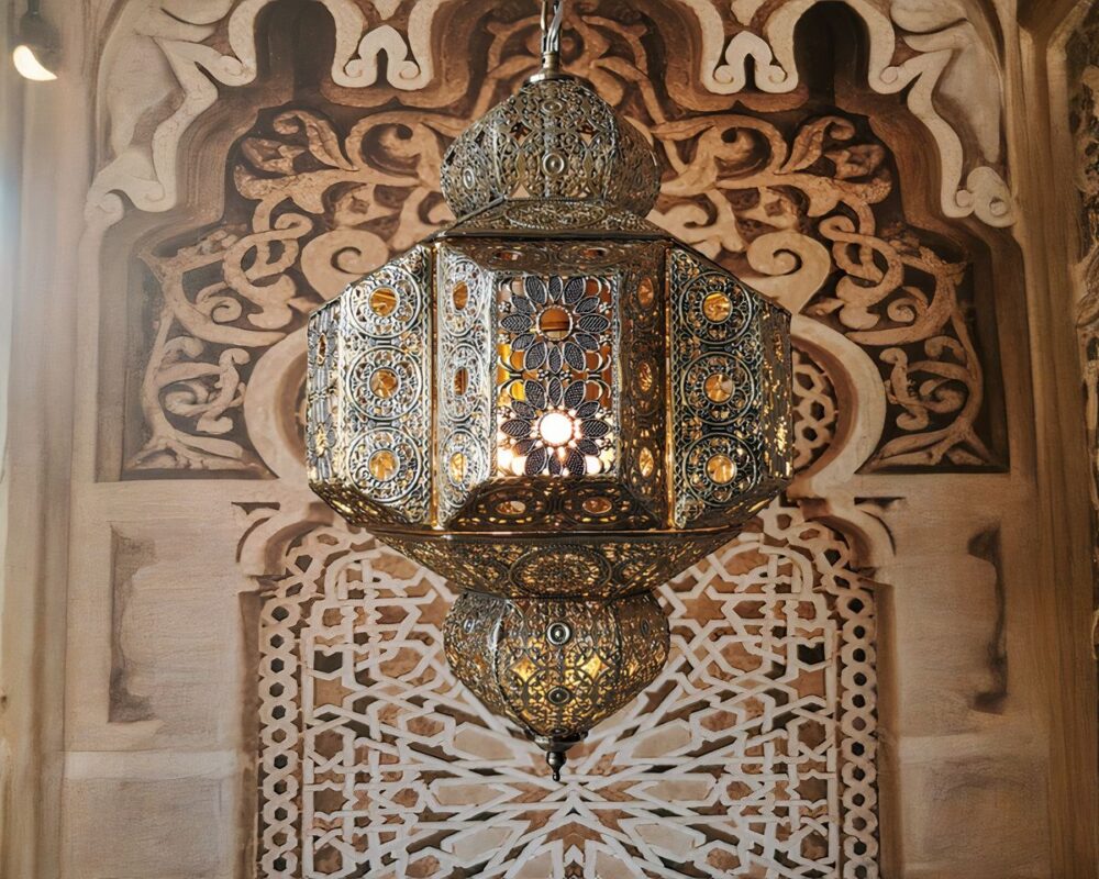 On voit un lustre marocain suspendu en métal vieilli de couleur doré virant sur le bronze. Il est en forme de lanterne traditionnelle en métal ajouré laissant voir que l'ampoule est allumée. A l'arrière, un mur avec des motifs orientaux de style arabesque marron et beige.