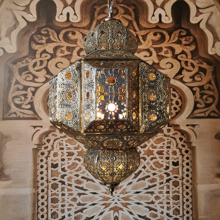 On voit un lustre marocain suspendu en métal vieilli de couleur doré virant sur le bronze. Il est en forme de lanterne traditionnelle en métal ajouré laissant voir que l'ampoule est allumée. A l'arrière, un mur avec des motifs orientaux de style arabesque marron et beige.