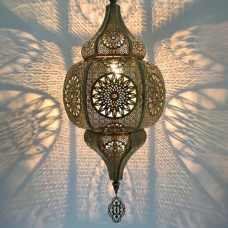 On voit un lustre marocain suspendu style vintage allumé. Son métal ajouré permet de diffuser des jeux de lumière sur le mur derrière. Il a la forme d'une lanterne vintage de couleur dorée.