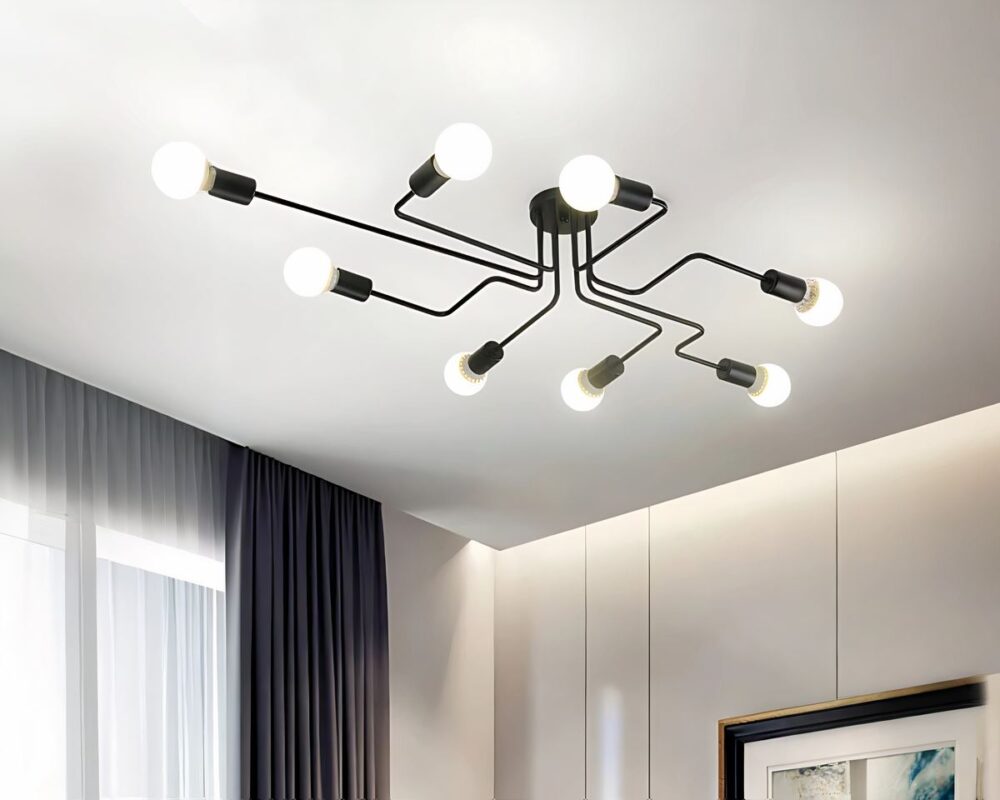 Lustre moderne à 8 branches au style industriel allumé et fixé sur un plafond blanc. Les 8 tiges coudées noires sont fixées sur un socle rond noir au centre. A chaque extrémité des tiges, une ampoule est allumée. Dans la pièce, on aperçoit à droite un mur blanc avec le coin d'un cadre. A gauche une fenêtre avec voilage blanc et rideau gris plissé sur la droite de la fenêtre.