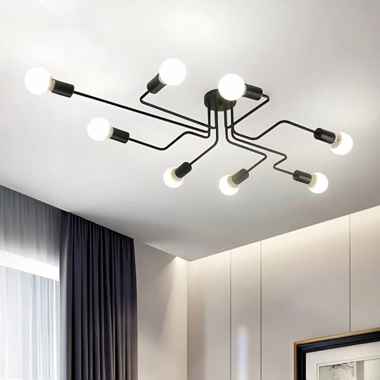 Lustre moderne à 8 branches au style industriel allumé et fixé sur un plafond blanc. Les 8 tiges coudées noires sont fixées sur un socle rond noir au centre. A chaque extrémité des tiges, une ampoule est allumée. Dans la pièce, on aperçoit à droite un mur blanc avec le coin d'un cadre. A gauche une fenêtre avec voilage blanc et rideau gris plissé sur la droite de la fenêtre.