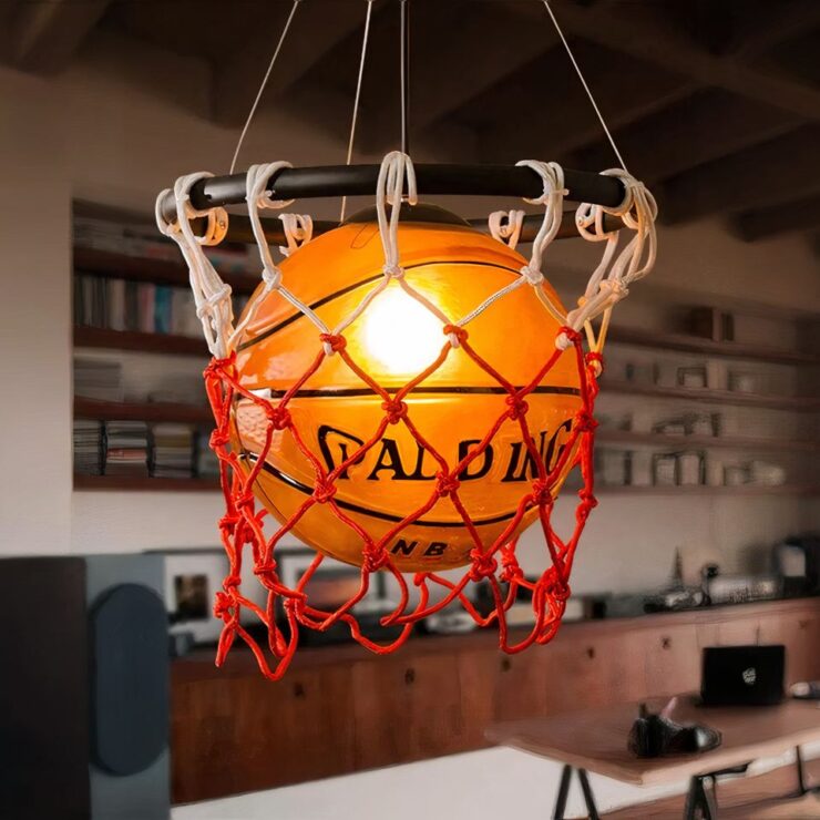 Lustre moderne en forme de ballon de basket allumé de couleur orange suspendu dans son panier filet rouge et blanc avec un anneau noir. A l'arrière on aperçoit un décor flouté avec une table en bois, un meuble bas long en bois et des étagères au mur.