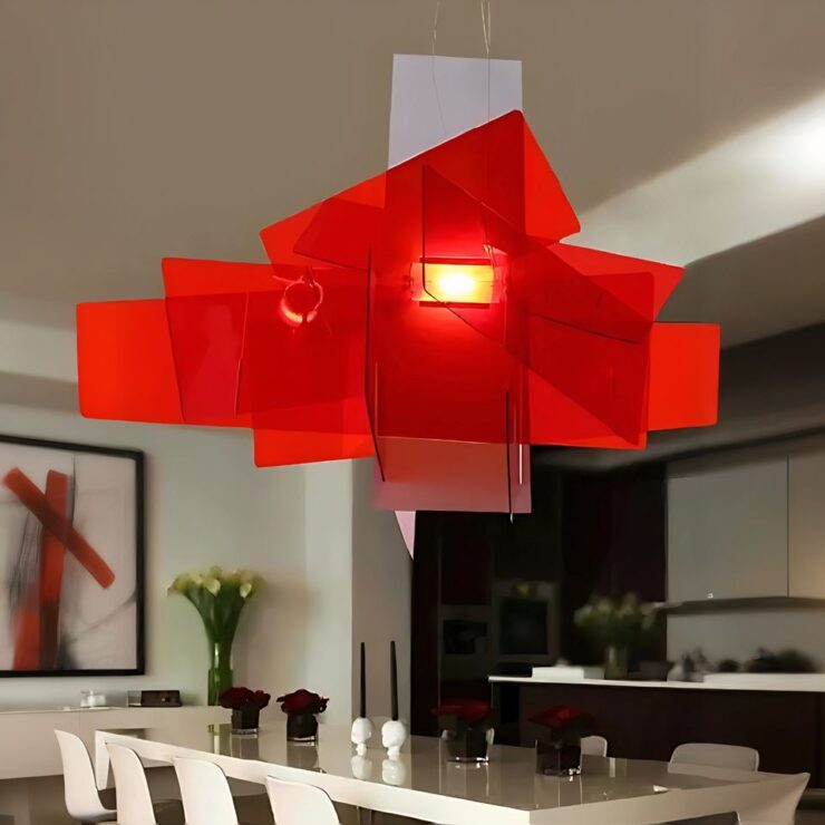 Lustre moderne inspiration big bang de foscarini de couleur rouge composé de plusieurs lames en acrylique rouge. Suspendu et allumé avec une ampoule au centre des lames. Dans la pièce, on aperçoit une grande table rectangulaire avec des chaises blanches autour. Sur la droite, un mur avec des étagères marron. Sur la gauche, un mur blanc avec un grand tableau.