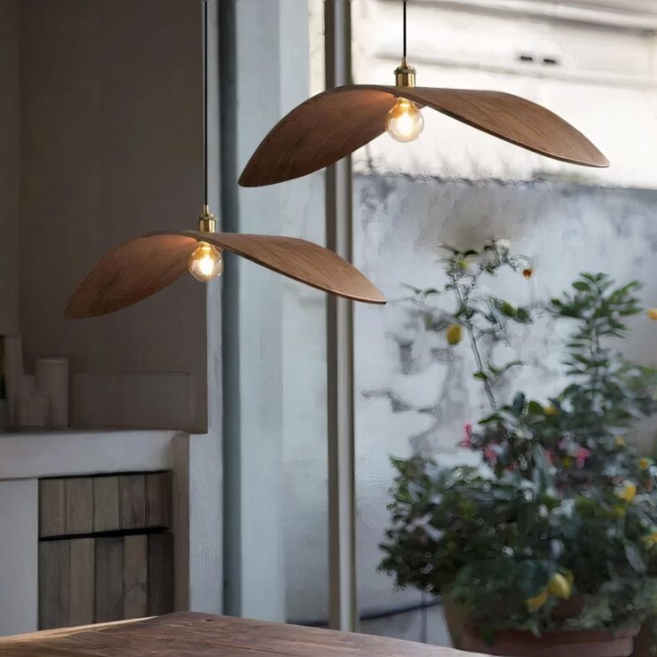 On voit deux lustres naturels en forme de feuille en bois servant d'abat-jour suspendu côte à côte. Leur ampoule est allumée. Ils sont fixés au-dessus d'une table en bois. Au fond, on peut voir une grande baie vitrée avec des plantes vertes en pot.