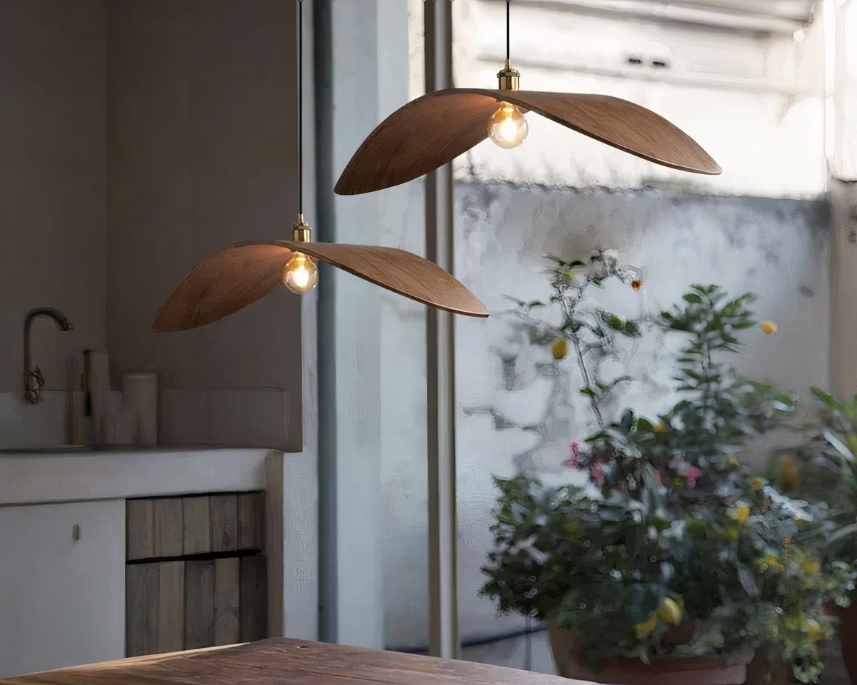 On voit deux lustres naturels en forme de feuille en bois servant d'abat-jour suspendu côte à côte. Leur ampoule est allumée. Ils sont fixés au-dessus d'une table en bois. Au fond, on peut voir une grande baie vitrée avec des plantes vertes en pot.