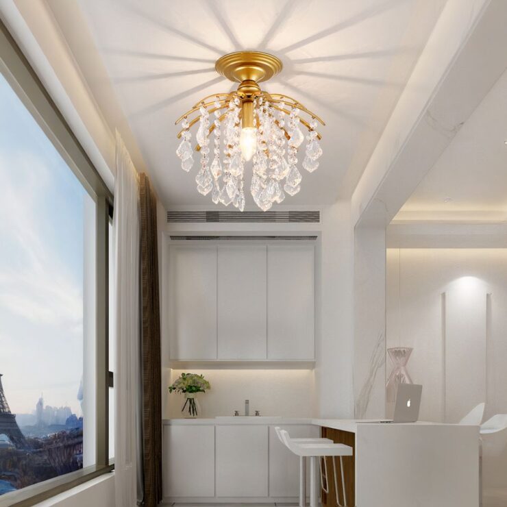 Un lustre baroque élégant avec cristaux suspendus, d’une couleur doré et décoré de cristaux transparents qui reflètent la lumière sur le plafond, créant des motifs lumineux. Il est installé dans une pièce moderne et lumineuse avec de grandes fenêtres et des meubles blancs.