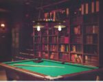 Lustre billard deux abat-jours suspendu au-dessus d'un grand billard en bois, avec une queue posée sur le tapis de jeu du billard.