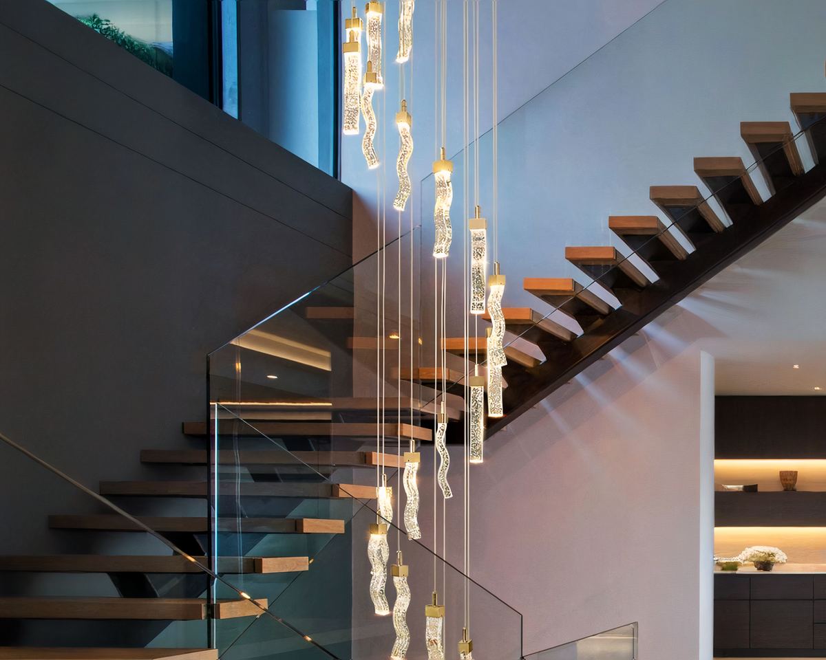Lustre de cage d'escalier vagues de cristal accrochée au plafond et mettant en valeur une montée d'escalier avec un garde corps en verre transparent. La lumière est tamisée, ce qui met en valeur le lustre.