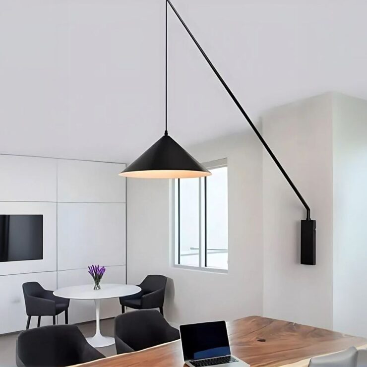 Lustre mural noir orientable fixé sur un mur blanc, entre deux fenetres est orienté au-dessus d'une table en bois sur laquelle est posé un ordinateur. En arrière-plan, on aperçoit une table ronde et blanche avec deux fauteuils noirs.