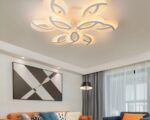 Le lustre est le format XXL, il est allumé, il est accroché au plafond, le lustre est en forme de pétales de fleur. IL se trouve dans un salon composé de deux canapés, un marron et un autre bleu. Le marron est un grand canapé avec plusieurs coussins. Le bleu a un coussin marron, il est situé au fond de la pièce devant une grande baie vitrée avec des grands voilages. Au milieu des canapés se trouve une table basse avec un vase en son centre.