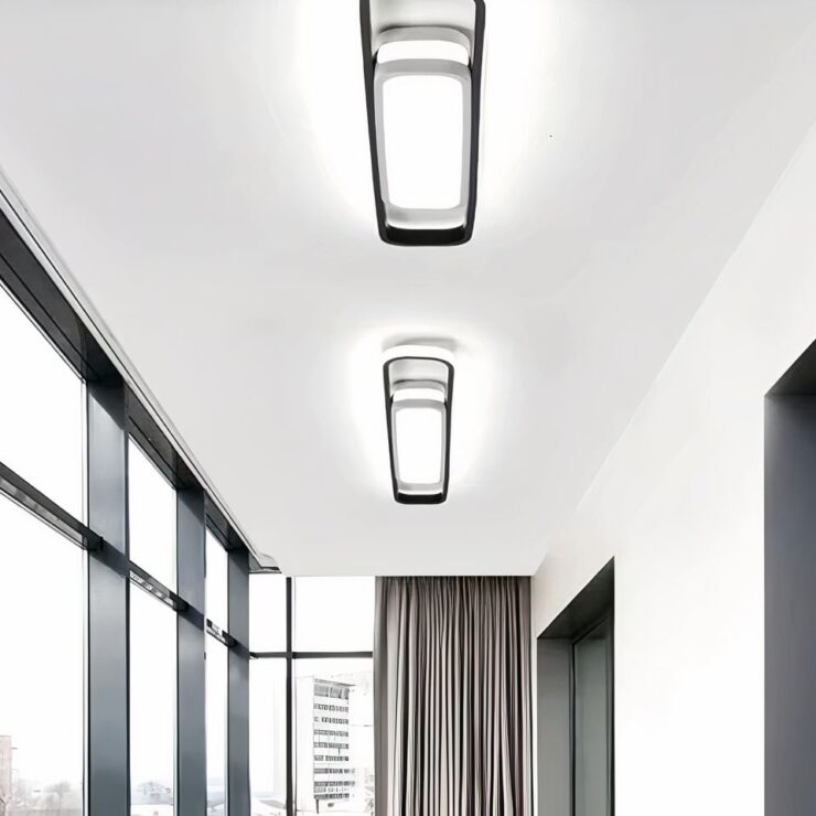 Lustre XXL plat, moderne, suspendu, les lumières sont allumés. Le lustre se trouve dans un grand couloir, entouré de grandes baies vitrés au fond on voit des rideaux gris.