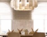 Lustre XXL , allumé, suspendu au plafond. Il est rond et il est composé de plusieurs sortes de tuiles. Il est suspendu au-dessus d’une table, au fond on peut voir deux grandes fenêtres en arc de cercle.
