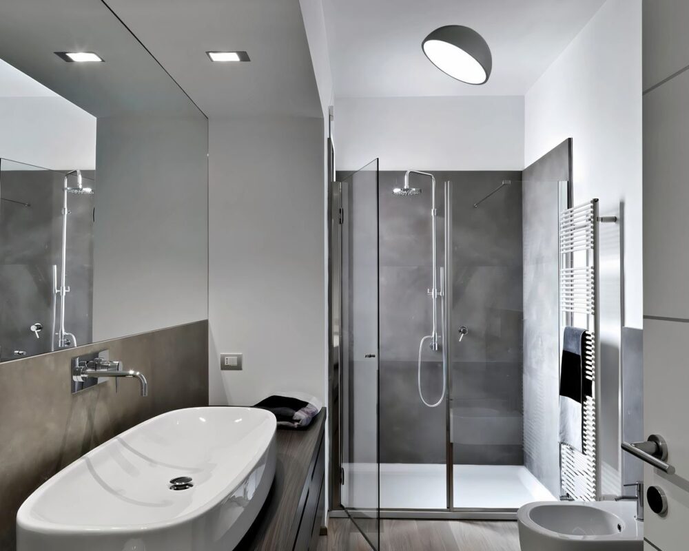 Lustre pour salle de bain en forme de demi-cercle, fixé au plafond. Ce luminaire moderne diffuse une lumière douce et homogène, éclairant élégamment l'espace avec son design épuré et fonctionnel, parfaitement intégré dans une salle de bain contemporaine aux tons gris et blancs.