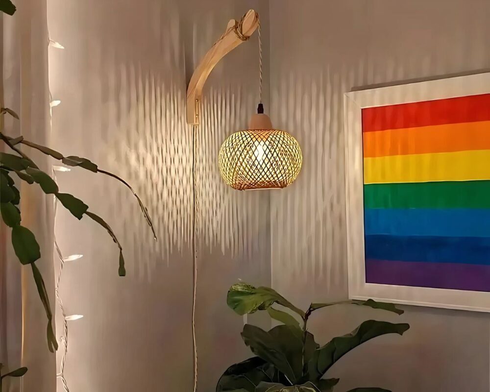 Applique lustre lanterne en bambou accrochée à un mur beige et allumée. On voit un cadre multicolore accroché au mur.