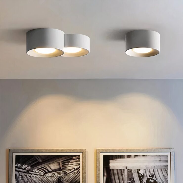 Un plafonnier LED cylindrique en saillie design minimaliste contemporain au plafond blanc, avec trois modules ronds gris éclairant doucement le mur au-dessus de cadres.