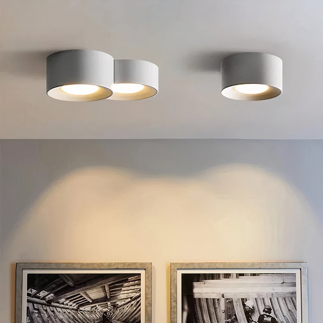 Un plafonnier LED cylindrique en saillie design minimaliste contemporain au plafond blanc, avec trois modules ronds gris éclairant doucement le mur au-dessus de cadres.