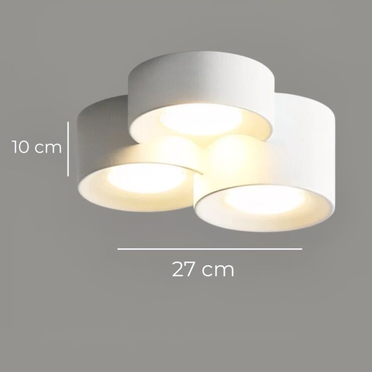 Un plafonnier LED cylindrique en saillie design minimaliste contemporain au plafond blanc, avec trois modules ronds gris éclairant doucement le mur au-dessus de cadres.
