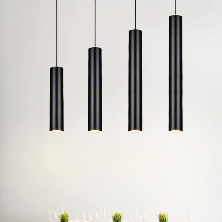 Quatre suspensions tube verticale design épuré moderne noires, longues et fines, pendent au plafond dans une pièce lumineuse au mur blanc.