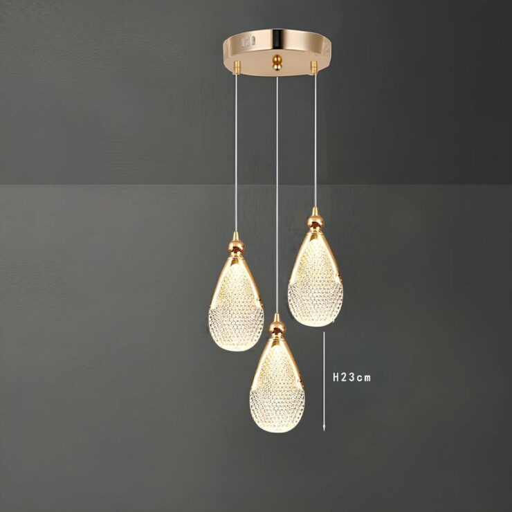 Un lustre goutte d’eau en acrylique doré suspendu au plafond, avec une forme de goutte brillante et un fond gris simple.