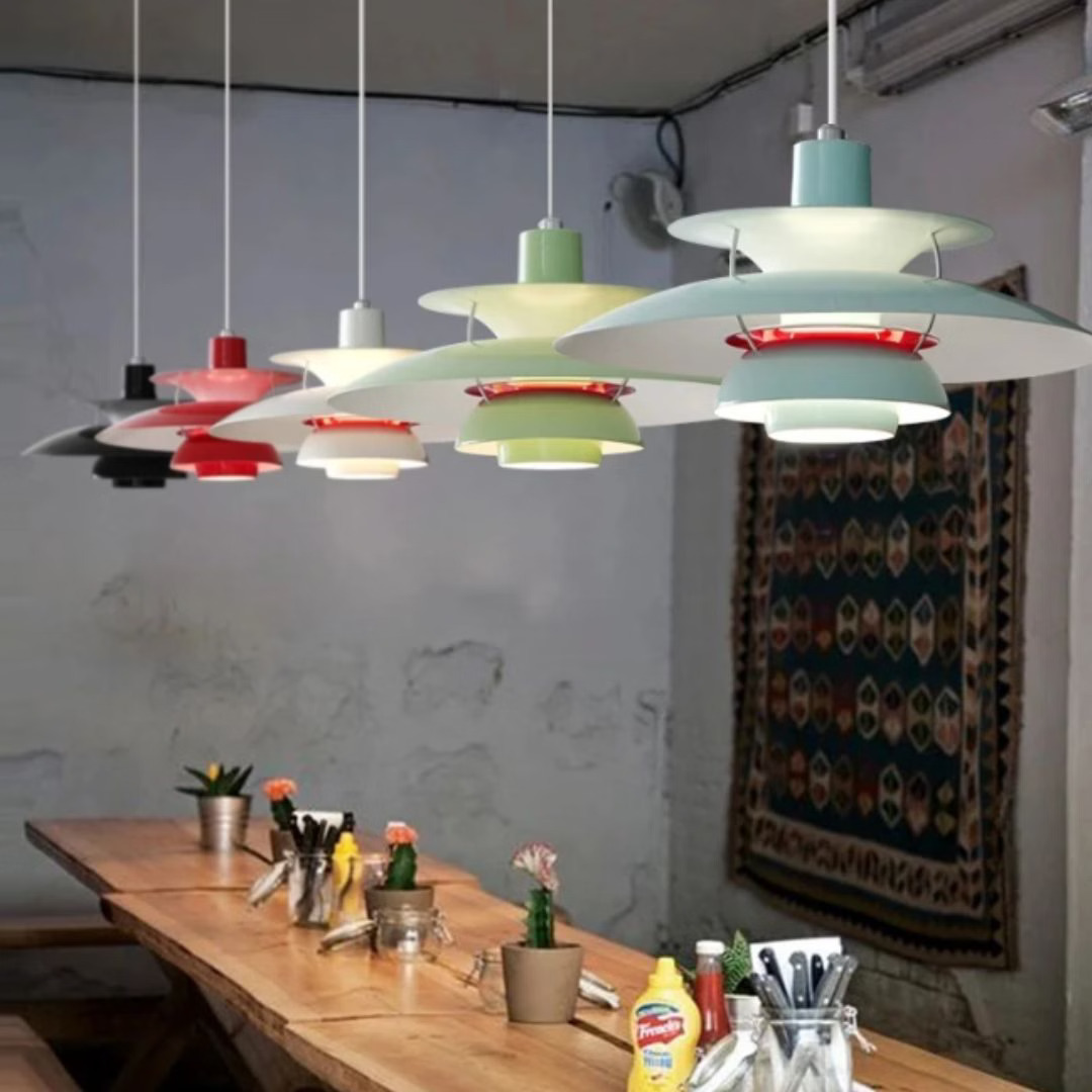 Plusieurs suspension design danois à abat-jour multicouches colorées pendent au-dessus d'une grande table en bois, dans une pièce au mur gris.